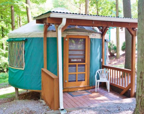 A green yurt with a wooden deck and a white chair, surrounded by trees in a serene outdoor setting.